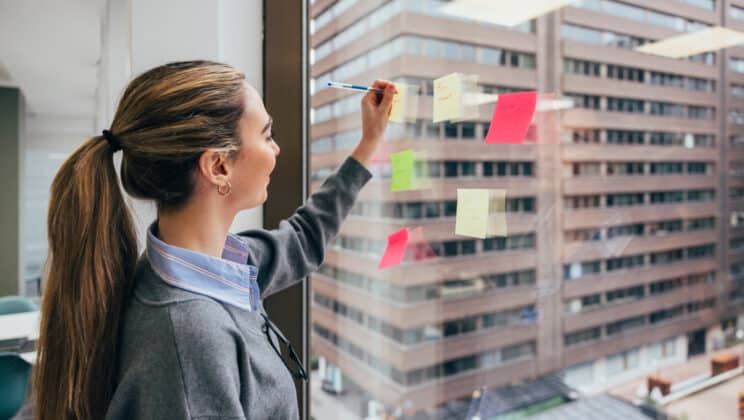 A focused young woman arranges colorful sticky notes on a glass window in a high-rise office building, overlooking an urban skyline.